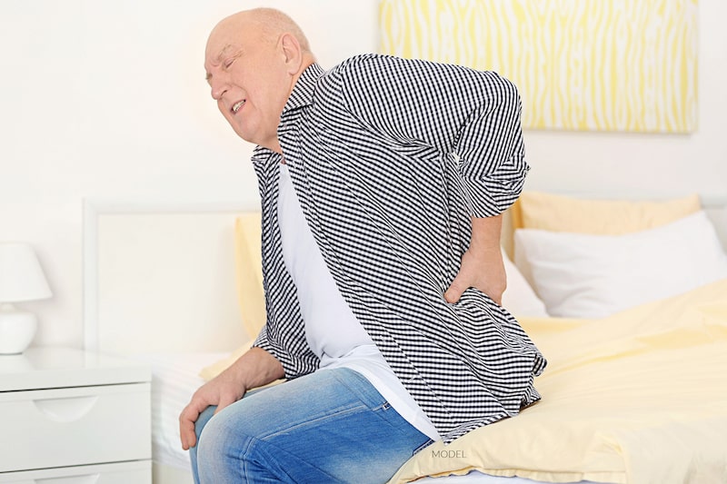 Senior man sitting on a couch and holding his lower back in pain.